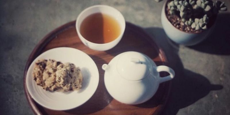 A white cup of amber tea and a white teapot on a wooden tray, beside a small succulent plant in sunlight