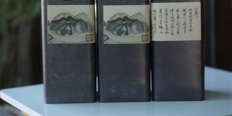 Three dark tea tins with painted mountain-and-river labels, set against a blurred garden window