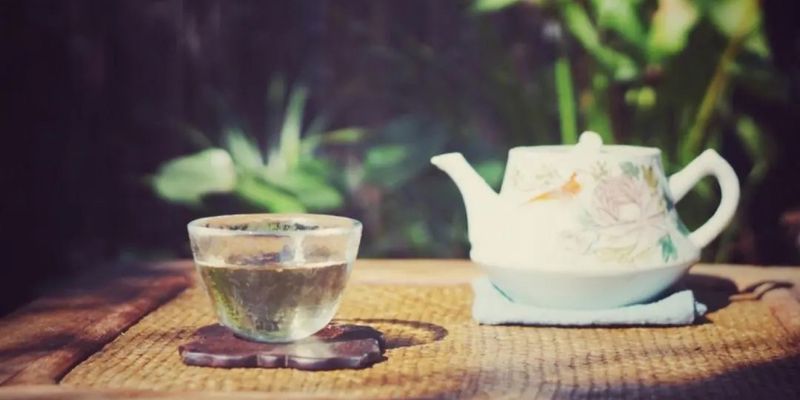 A glass cup of pale jasmine tea beside a floral porcelain teapot, set on a woven mat in dappled garden light