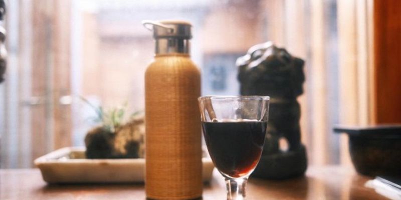 A glass of dark tea beside a woven bamboo flask, with a small stone figure blurred in the background