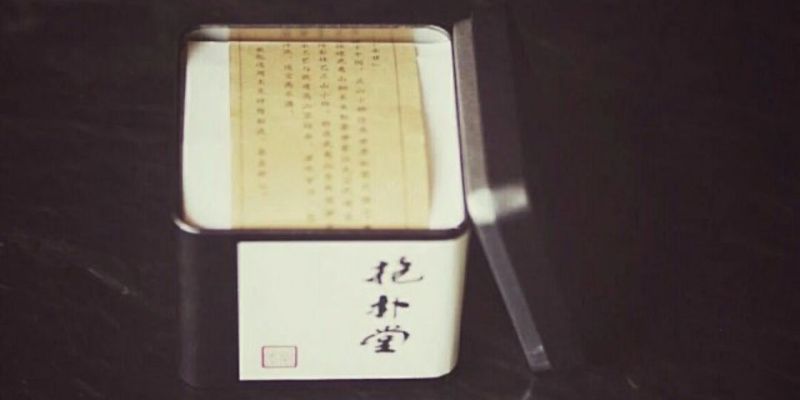 An open square tea tin showing a printed paper insert, lid resting against the tin