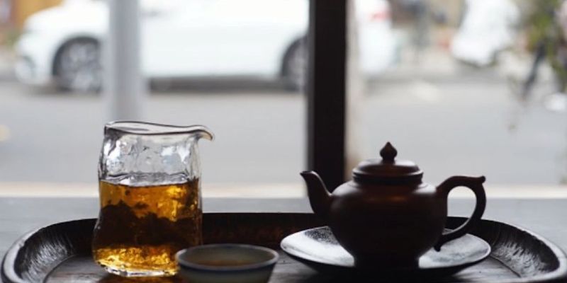 A glass pitcher of amber tea and a small dark teapot on a tray, with a street scene blurred through a window behind