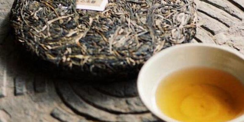 A loose-leaf raw puer cake beside a small cup of golden tea, resting on a carved stone surface