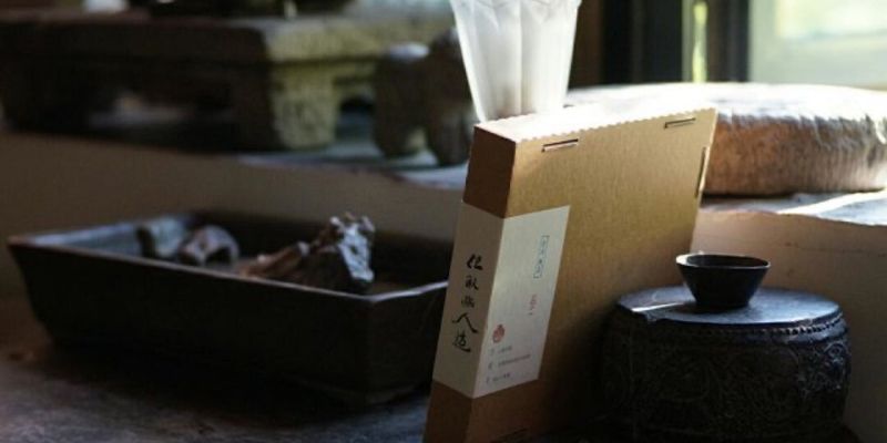 A dim, sunlit tea room with a packaged tea box on a stone table, a black ceramic jar and cup beside it