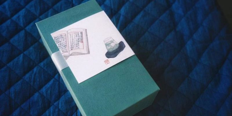 A teal gift box with an illustration of an open book and a glass jar, resting on a quilted blue fabric