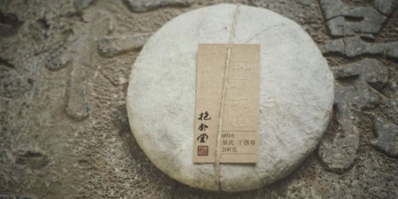 A round raw puer cake wrapped in twine with a paper label, resting on a weathered stone carving