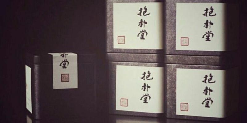 Several dark tea tins stacked together, one with its paper wrap still loose, against a dark background