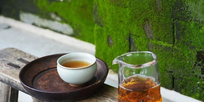A white cup of amber tea and a glass pitcher of the same tea, set on a dark wooden bench against a mossy stone wall