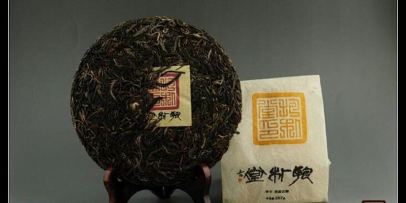 A round raw puer cake on a wooden display stand next to its paper wrapping, studio lit against a grey background