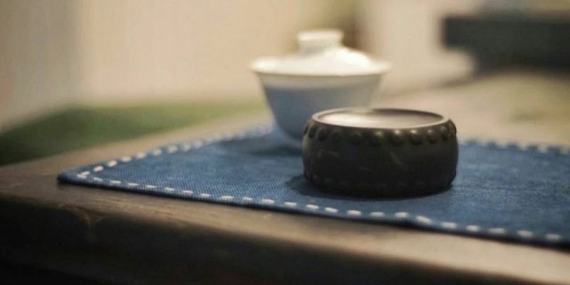 A small dark drum-shaped inkstone on a stitched blue cloth, beside a blurred white gaiwan