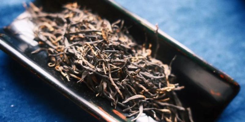 Loose dark, golden-tipped black tea leaves spread on a black tray against a blue fabric backdrop