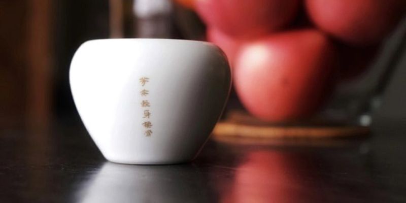 A small white porcelain cup standing on a dark wooden table, fruit blurred in the background